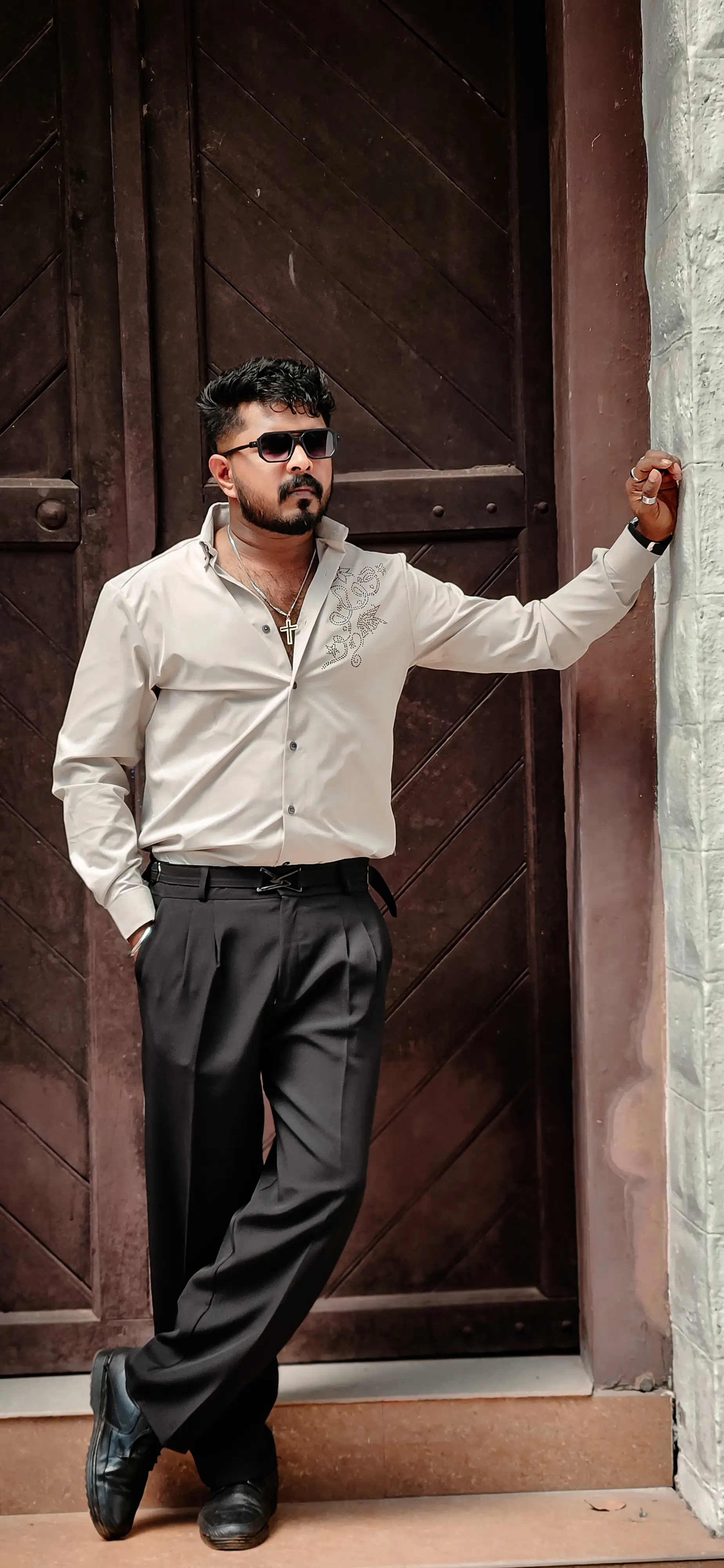 alt="Ebin abraham in a cream shirt and sunglasses posing by a dark wooden door.''